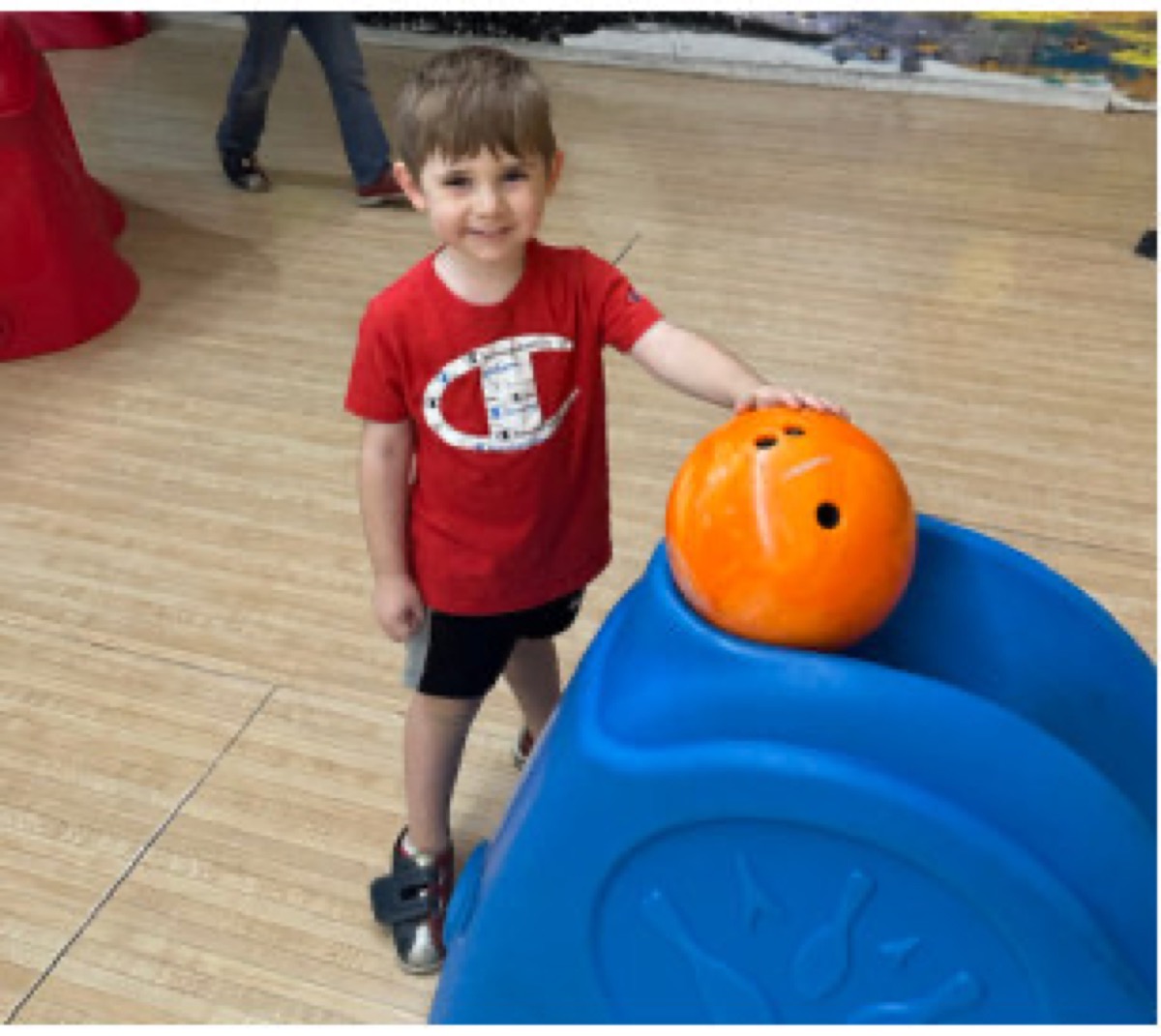 A young bowler smiling with an orange bowling ball on the ball ramp